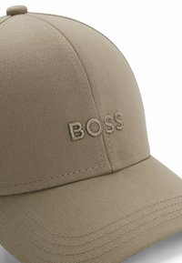 Beige baseball cap made of textured fabric, featuring embroidered "BOSS" logo on the front, with stitched accents along the brim.