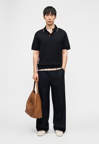 Άνδρας με μαύρο πόλο πουκάμισο και φαρδιά μαύρη παντελόνα κρατάει μεγάλη καφέ δερμάτινη τσάντα, στέκεται μπροστά σε απλό λευκό φόντο.