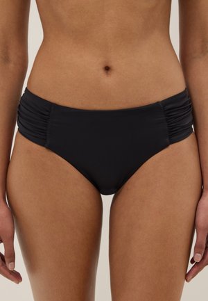 Vrouw met zwarte bikinibroekjes met gerimpelde zijkanten, gefotografeerd van taille tot halverwege de dij tegen een neutrale achtergrond.