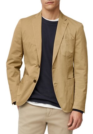 Hombre con blazer beige sobre suéter azul marino y camisa blanca, combinado con pantalones beige, recortado a la altura del pecho y por encima de las rodillas.