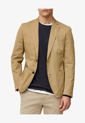 Mężczyzna w beżowej marynarce na granatowym swetrze i białej koszuli, w beżowych spodniach, zdjęcie od klatki piersiowej do ponad kolan.