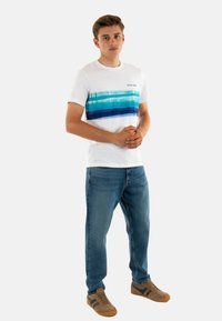 Camiseta blanca con un diseño de rayas horizontales azul-verdosas, combinada con jeans azules y zapatillas marrones. Diseño informal con un corte relajado.