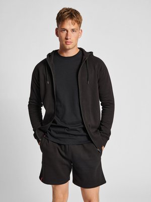 Jeune homme portant un sweat à capuche noir zippé, un t-shirt noir et un short noir, debout les mains dans les poches devant un fond gris uni.