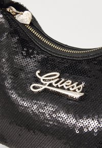 Czarna kopertówka z cekinami o błyszczącej fakturze, z detalami w kolorze złotym oraz wyraźnym białym akcentem z logo "Guess". Posiada charms w kształcie serca.