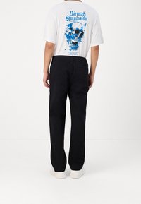 Vit t-shirt med blått dödskallemotiv på ryggen, kombinerad med svarta byxor med avslappnad passform och vita sneakers, vilket ger en avslappnad stil.