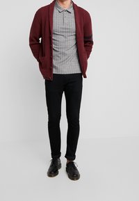 Cardigan rosso in maglia con bottoni sopra una polo grigia a righe. Jeans neri slim fit e scarpe nere con lacci completano il look.