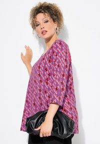 Top rosso e blu con motivo, dalla vestibilità ampia e scollatura larga, abbinato a una pochette nera con texture. Il modello tiene la pochette con una mano.