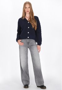 Granatowy sweter z dzianiny o fakturowym wzorze, z dekoltem w serek oraz pięcioma okrągłymi guzikami, zestawiony z jasnoszarymi spodniami o szerokich nogawkach i czarnymi botkami.