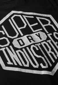 Camiseta de algodón negra con letras blancas prominentes y texturizadas en forma hexagonal, que dicen "SUPER DRY INDUSTRIES."