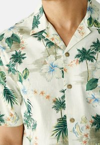 Koszula z krótkim rękawem z lekkiego materiału, z tropikalnym wzorem zawierającym zielone liście palmowe i niebieskie kwiaty hibiskusa, z zapięciem na guziki i beżowymi guzikami.
