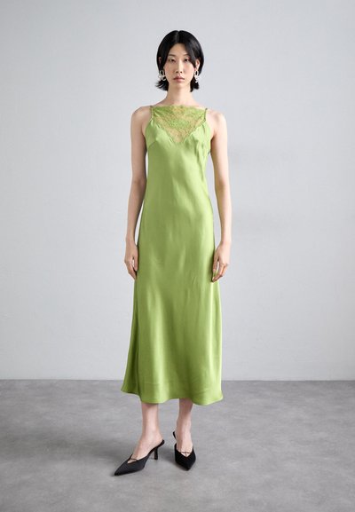 Robe de chambre en satin vert avec finition en dentelle au col ; design longue, silhouette adaptée, associée à des mules noires à talons pointus.