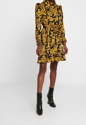Femme portant une mini-robe à manches longues noire et jaune à fleurs avec une ceinture à la taille et des bottines noires texturées.