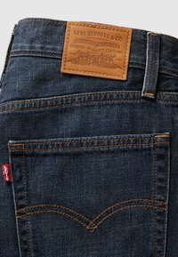 Donkerblauwe denim jeans met oranje stiksels, voorzien van een bruine leren patch met een reliëflogo en twee achterzakken met gebogen naden.