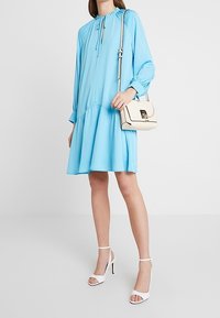 Ljusblå, långärmad klänning med plisserad kjol och samlad halsringning. Kombinerad med en gräddfärgad handväska och vita högklackade sandaler.