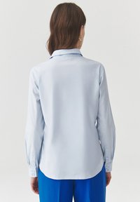 Camisa de manga larga azul clara con cuello, puños abotonados y una textura suave. La parte trasera tiene un bajo ligeramente curvado.