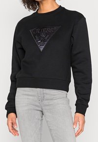 Sudadera negra con un diseño recortado, que presenta un parche triangular texturizado con el logo "GUESS". Puños y dobladillo acanalados, material suave.