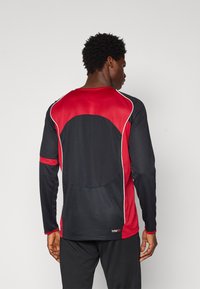 Tricou sport cu mâneci lungi, având un corp principal negru, accente din plasă roșie și cusături albe. Include un logo pe partea de jos a spatelui.