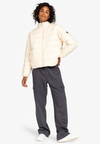 Puffig, krämfärgad jacka med hög krage, dragkedjestängning och textilstruktur. Bärs med grå cargobyxor och vita sneakers.