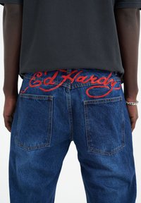 Jeans de mezclilla azul con el logotipo bordado en rojo "Ed Hardy" en la pretina, que cuentan con dos bolsillos traseros y un diseño de pierna recta.