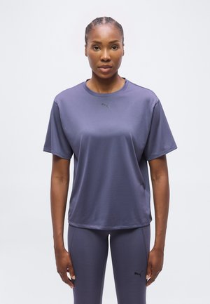 Femme portant un t-shirt de sport Puma violet foncé assorti à un legging, debout devant un fond clair uni.