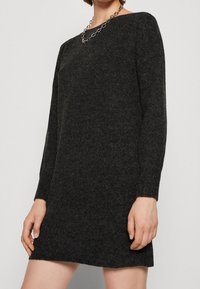 Osoba má na sobě tmavě šedé pletené šaty s dlouhými rukávy a stříbrný řetízek, pózuje s jednou rukou opřenou o bok.