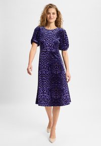 Lila Samtkleid mit Leopardenmuster, kurzen Puffärmeln und schwingendem Saum, kombiniert mit hellen spitzen Absätzen.