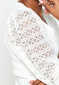 Biała bluzka z teksturowanej tkaniny, z misternymi koronkowymi rękawami prezentującymi diamentowy wzór oraz luźnym krojem.