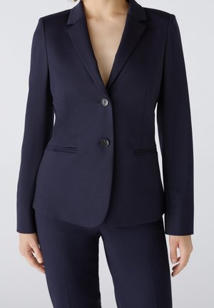Personne portant un blazer bleu marine foncé sur mesure avec deux boutons et un pantalon assorti, debout devant un fond clair.