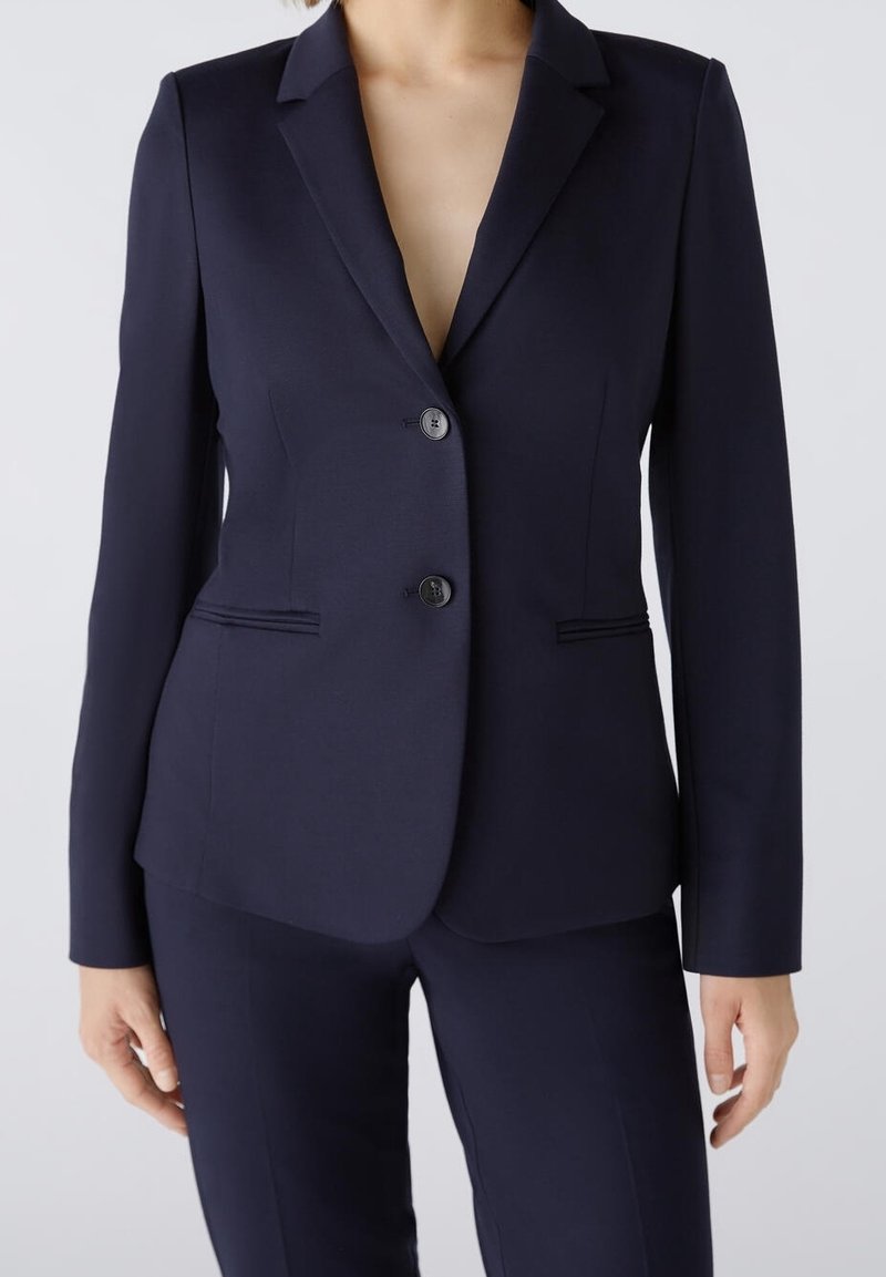 Personne portant un blazer bleu marine foncé sur mesure avec deux boutons et un pantalon assorti, debout devant un fond clair.