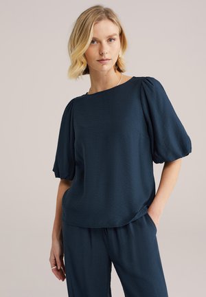 Femme blonde portant un chemisier bleu marine à manches bouffantes et un pantalon assorti, debout avec une main dans la poche, devant un fond uni.