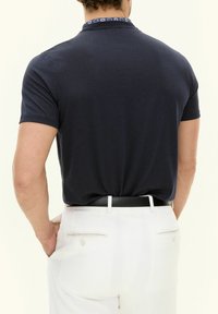 Short-sleeve navy polo shirt with a patterned collar, paired with white pants. Features a fitted design and a smooth knit texture.