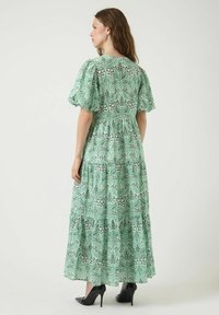 Robe maxi verte à fleurs avec manches bouffantes, design à volants, tissu texturé et détail dos avec une ouverture en forme de goutte, associée à des talons hauts noirs.
