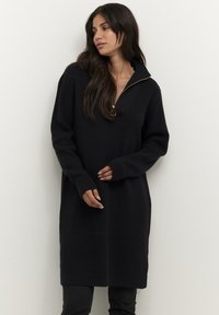 Robe en tricot noire avec un col zippé mi-haut. Elle présente des manches longues, des poignets côtelés et une coupe décontractée, arrivant au milieu de la cuisse. Surface texturée.