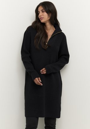 Robe en tricot noire avec un col zippé mi-haut. Elle présente des manches longues, des poignets côtelés et une coupe décontractée, arrivant au milieu de la cuisse. Surface texturée.