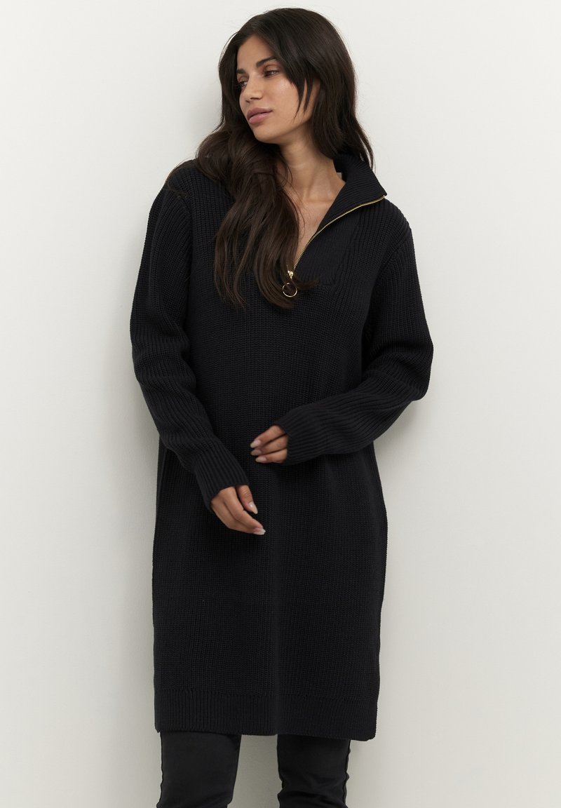 Robe en tricot noire avec un col zippé mi-haut. Elle présente des manches longues, des poignets côtelés et une coupe décontractée, arrivant au milieu de la cuisse. Surface texturée.