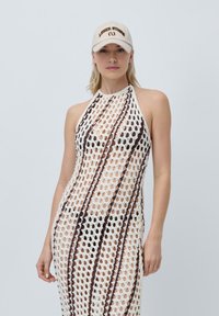 Robe halter au crochet de couleur blanche avec des rayures marron, présentant un design en maille ouverte. Associée à une casquette beige affichant "LEGER STUDIO" en marron foncé.