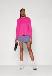Långärmad rosa träningströja med rund hals, i kombination med lila shorts. Modellen håller en svart vattenflaska och har på sig ljuslila sneakers.