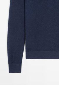 Manica lunga di un maglione blu navy e sezione del busto inferiore con polsini e orlo a coste su sfondo bianco.