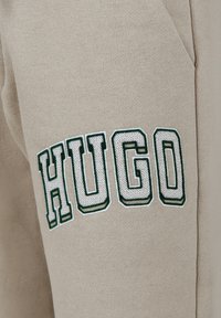 Tissu beige avec des lettres blanches entourées de vert épelant "HUGO" sur des vêtements, montrant des détails de texture et de couture sur une couture latérale.