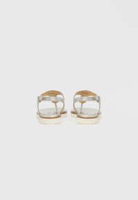 Αντηλιακά σανδάλια με ασημί γκλίτερ, επίπεδη, ανοιχτόχρωμη σόλα, λεπτά λουράκια και λεπτομέρεια με πόρπη. Οπίσθια όψη που δείχνει λείο πέλμα και λεπτή σχεδίαση.