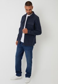 Marineblå uld skjorteblazer med to brystlommer, knaplukning og en afslappet pasform, parret med lyseblå denim og hvide sneakers.