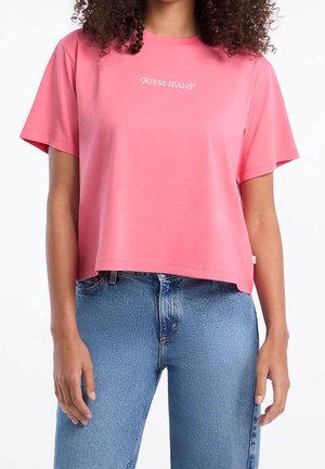 Kobieta w krótkim różowym T-shircie z napisem „GUESS JEANS”, w parze z wysokim stanem niebieskich dżinsów, stojąca z rozluźnionymi ramionami.