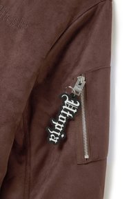 Giacca in finta pelle scamosciata marrone con ricamo "Utopia" e una tasca con zip argentata. Include un portachiavi nero con testo bianco attaccato alla zip.