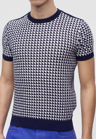 Homme portant une chemise en maille à manches courtes, à motif bleu marine et blanc, avec col et poignets bleu marine, associée à un pantalon bleu.