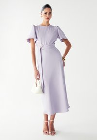 Lila midi obleka z naborki, z pasom na pasu in okrožno sponko. Manekenka drži belo geometrijsko torbico in nosi sandale s trakovi.