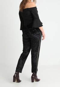 Svart off-shoulder topp med utsvängda ärmar, parat med svarta sammetbyxor med en glänsande sidos stripes, bärs med mörka klackboots.