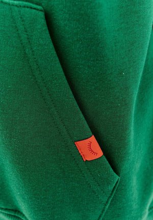 Zelená mikina s jemnou textúrou, ktorá má vrecko a malú červenú nášivku s grafickým detailom.