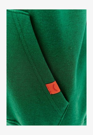 Zelená mikina s kapucí, vyrobená z měkkého materiálu, s kapsou a malou červenou nálepkou se šitým grafickým detailem.