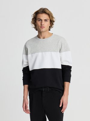 Giovane uomo con capelli biondi ondulati che indossa una felpa a maniche lunghe a blocchi di colore in grigio, bianco e nero, in piedi davanti a uno sfondo chiaro e uniforme.