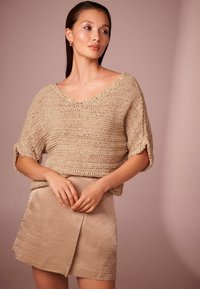 Haut en tricot beige avec un col en V et des manches roul�ées, associé à une jupe enveloppante beige clair ayant un léger éclat et une texture lisse.